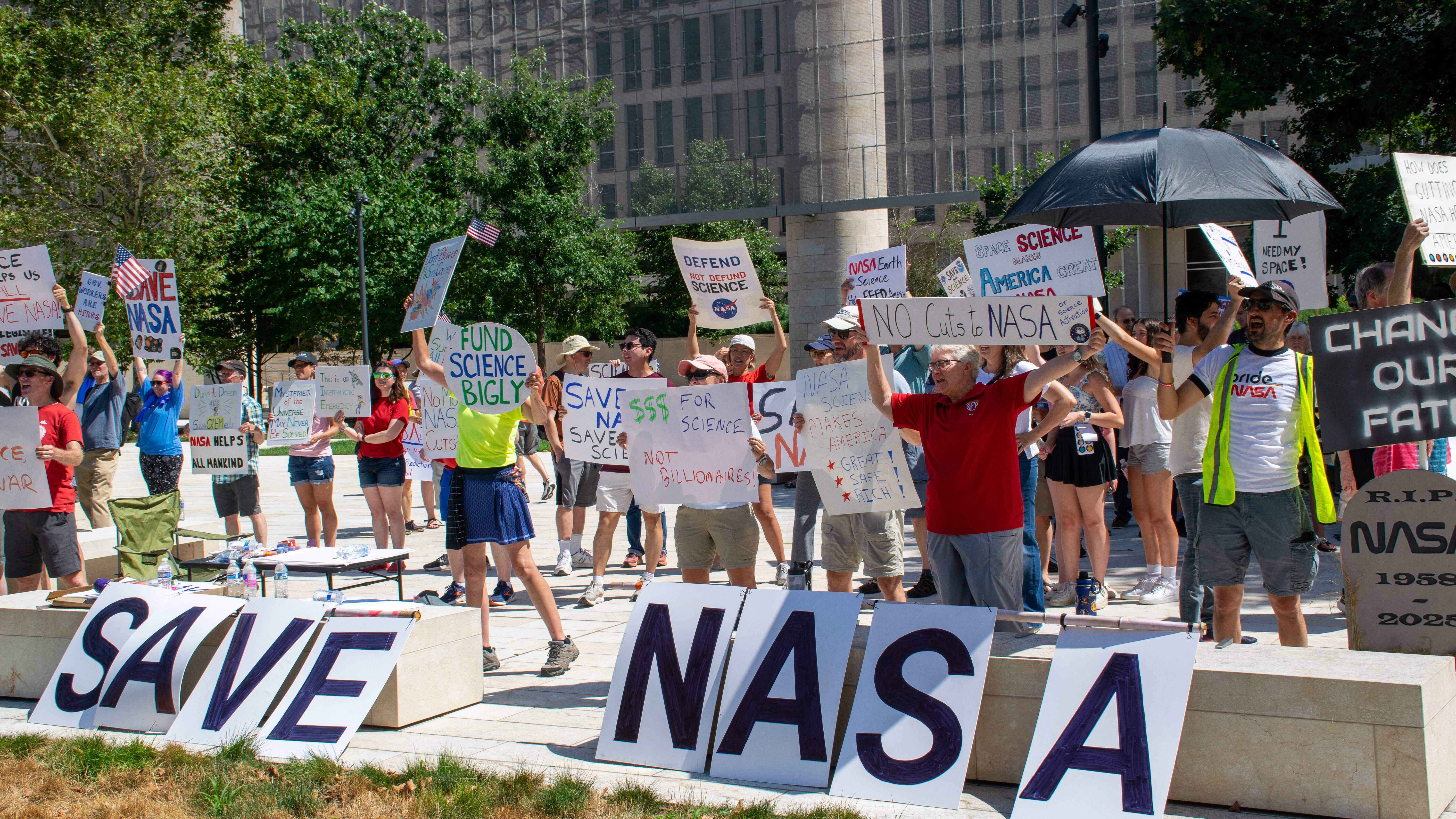 People hold signs outside on a sunny day in protest of NASA budget cuts, July 20, 2025.