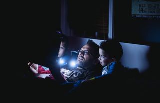 A man in bed with his two children, reading a bedtime story with a flashlight