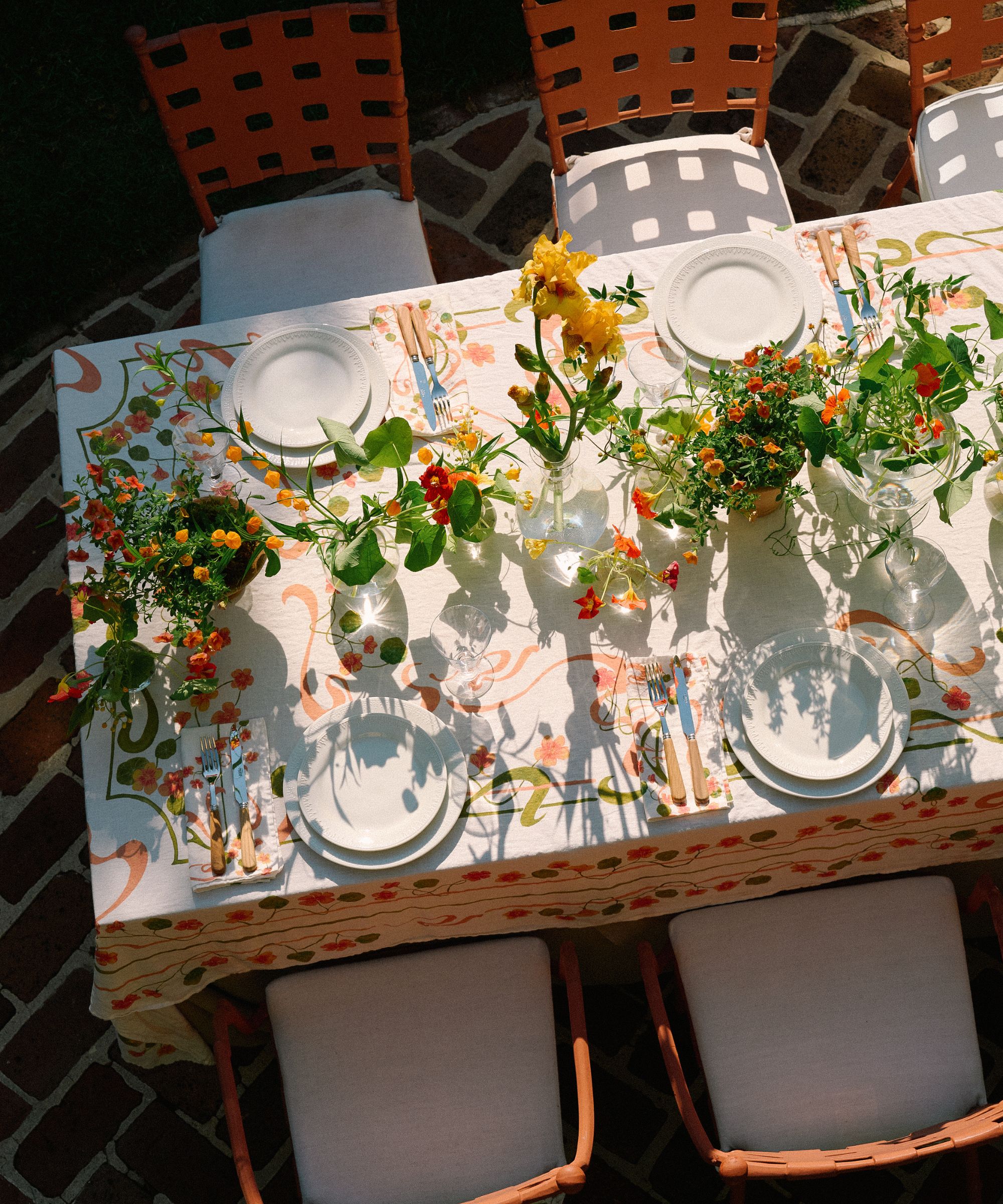 Outdoor dining table with orange metal chairs, a floral orange and white tablecloth, orange flowers in vases along the center of the table, white plates, and bamboo cutlery.