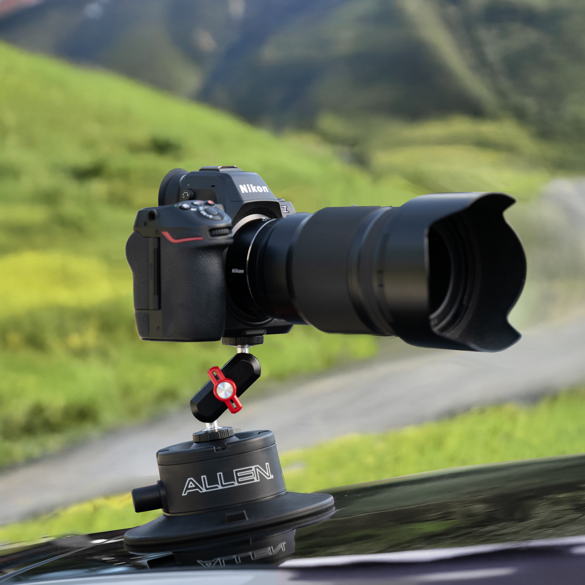 An Allen Smart Suction Snap Camera Mount is attached to a black car hood, supporting a Nikon mirrorless camera with a large lens against a backdrop of green hills.