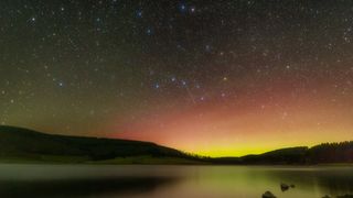 a starry night sky above a lake, with the silhouette of a mountain range extending from left to right across the background