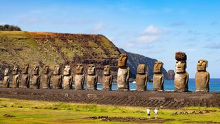 A couple stands next to a row of giant statues on Rapa Nui by the beach.