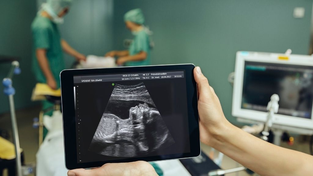 Hands holding up a screen with an ultrasound in front of a surgical table and team of doctors prepping for operation