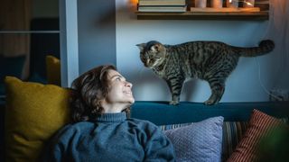 Woman sitting on a sofa looking up at her cat behind her who is standing on the back of the sofa