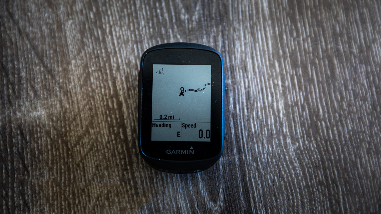 A Garmin Edge 130 Plus cycling computer on a wooden surface, with a basic map showing the current location and a route line, with 'heading' showing East and 'speed' showing 0.0mph