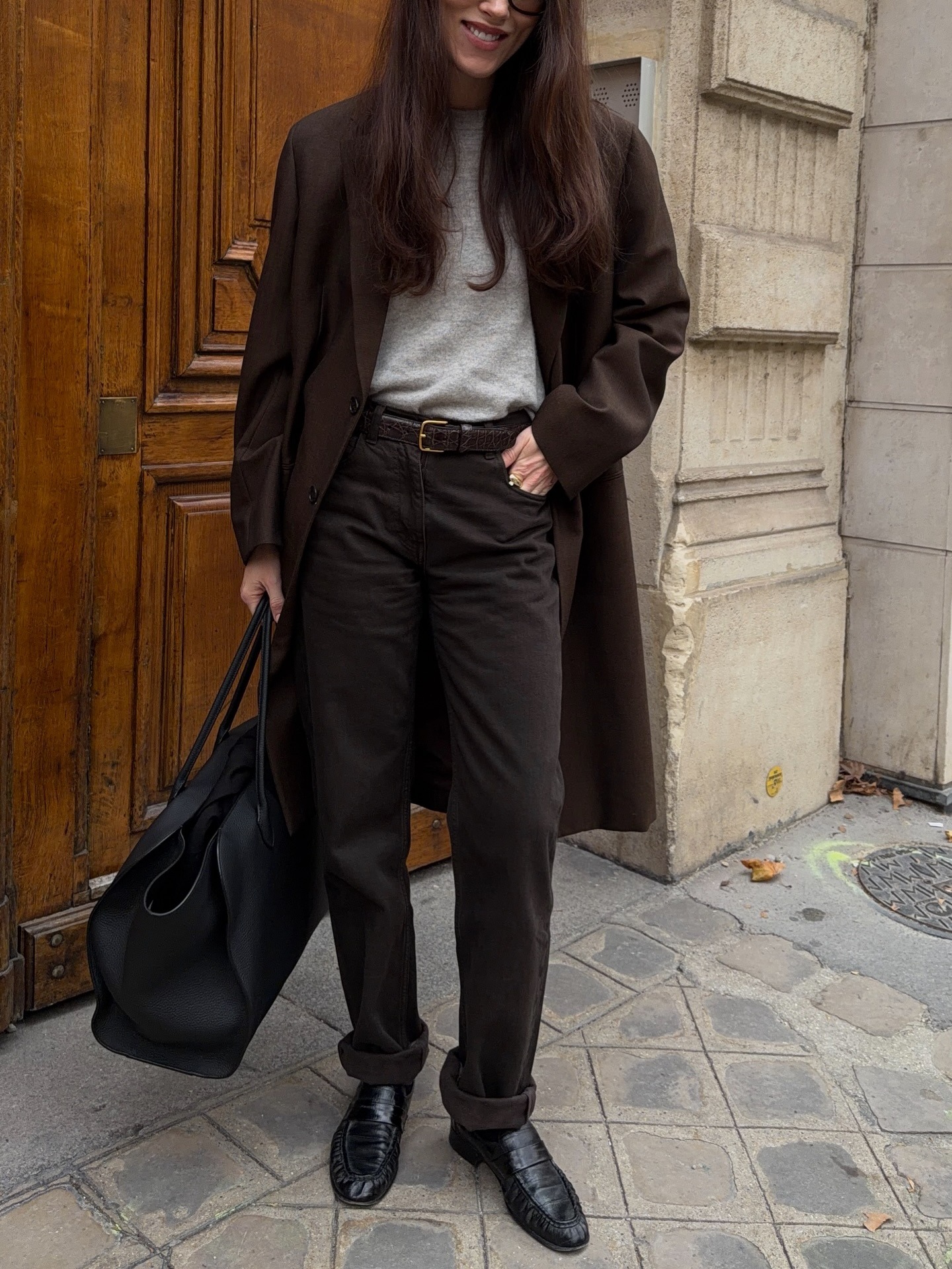 Annabel wears a brown coat, gray tee, brown jeans, black loafers, and a black duffel bag.