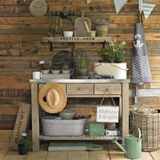 A potting bench in a wooden shed with flower pots, watering can, basket, seeds to sow, bunting and straw hat.