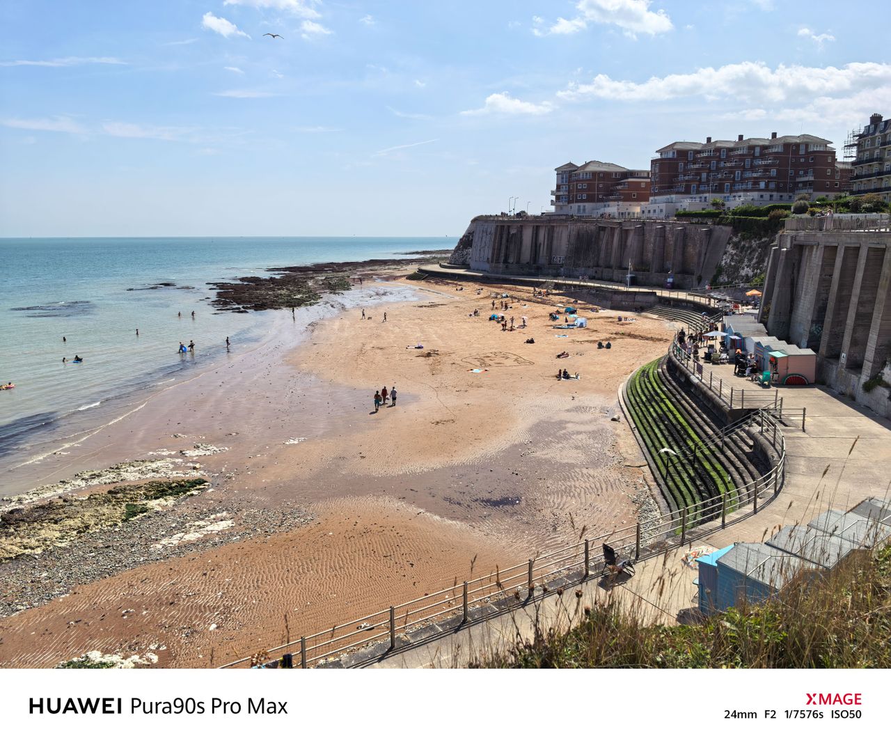 Sunny coastal beach at low tide, with people on the sand and seafront buildings above the cliffs.