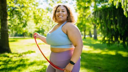 A woman in a sports bra and leggings stands outside with a resistance band in her hands, smiling at the camera. She has a fitness tracker on her wrist and there are leafy trees behind her. 