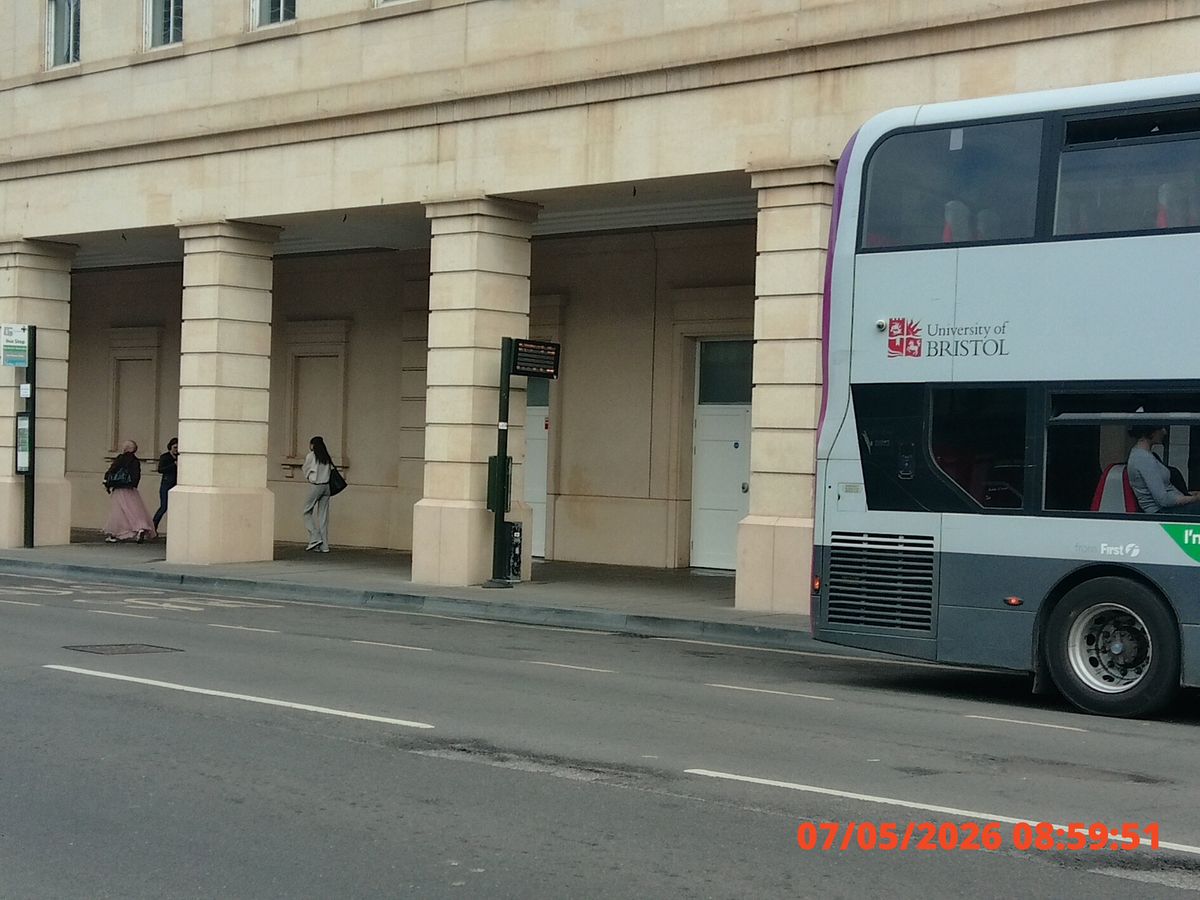City street scene with a red and white double-decker bus passing by a building with columns. Three people stand under the arches. Bus displays "University of Bristol"