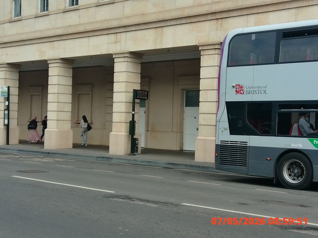 City street scene with a red and white double-decker bus passing by a building with columns. Three people stand under the arches. Bus displays "University of Bristol"