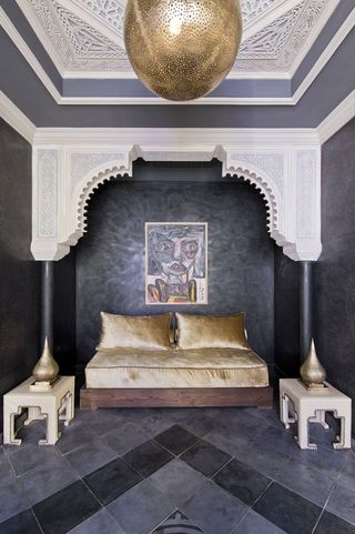A hotel sitting area with a gold fabric sofa, white side tables with gold ornaments, white arched roof, dark grey pillars, dark grey walls, a wall painting and square floor tiles in shades of grey.