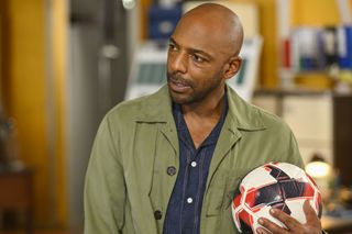 DI Mervin Wilson (Don Gilet) stands in police headquarters, wearing a blue shirt and a green jacket. He is clutching a football in his left hand.