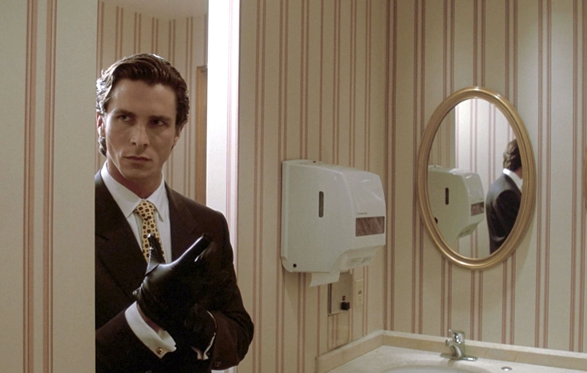 Patrick Bateman (Christian Bale) walks into the bathroom while wearing a black glove in American Psycho.