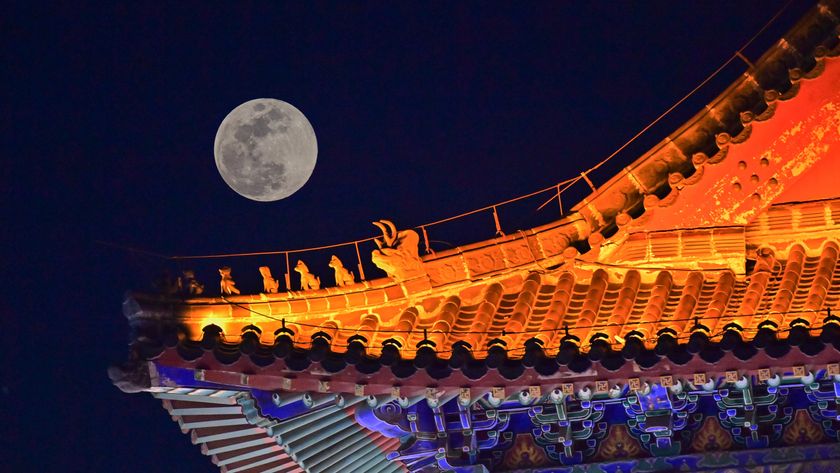 full moon on the left shining next to an ornate roof of a building illuminated by lights. 