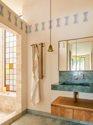 A beige powder room with baby blue, geometric tiles at 3/4 height. To the right is a custom, turquoise basin, with a wood cabinet beneath. The space is floored with a pale, turquoise and a pinky beige lite with a multi-coloured window to the left. In the middle hangs a small, bronze pendant light with vertical, bronze towel warmers behind.