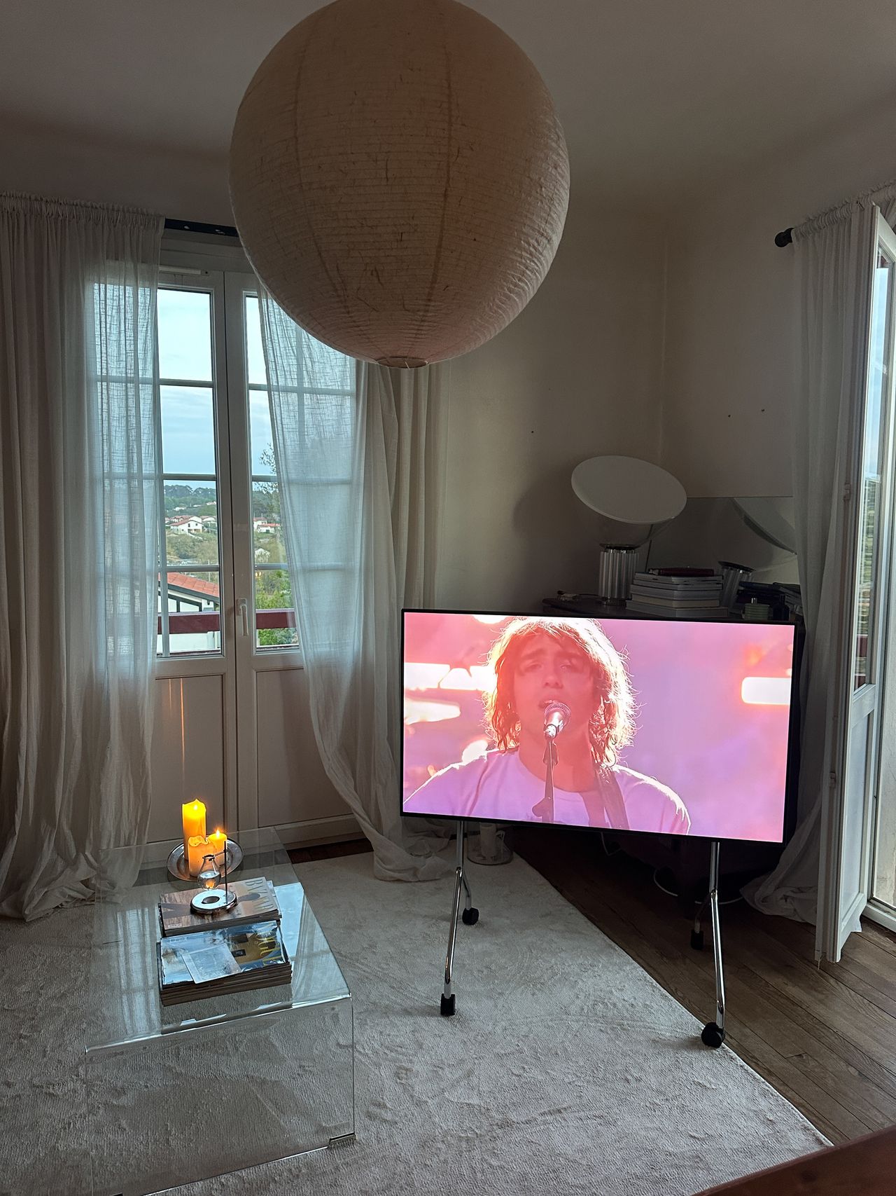 Image of a TV on a rolling TV stand in a whtie living room that has white curtains, a white rug, and a clear coffee table.