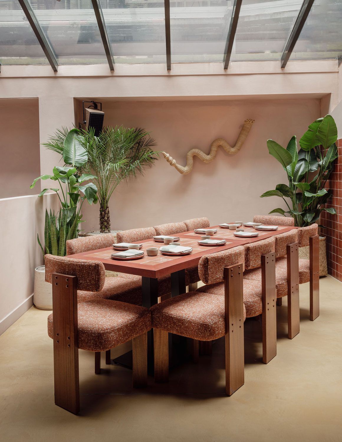 A private dining area in a restaurant features a long, terracotta-hued wooden table decorated with glazed tiles, upholstered wooden chairs in a fuzzy fabric, trailing plants, and a snake sculpture hanging from the wall.