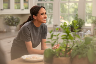 Meghan Markle wearing a gray t-shirt leaning on a kitchen counter and smiling