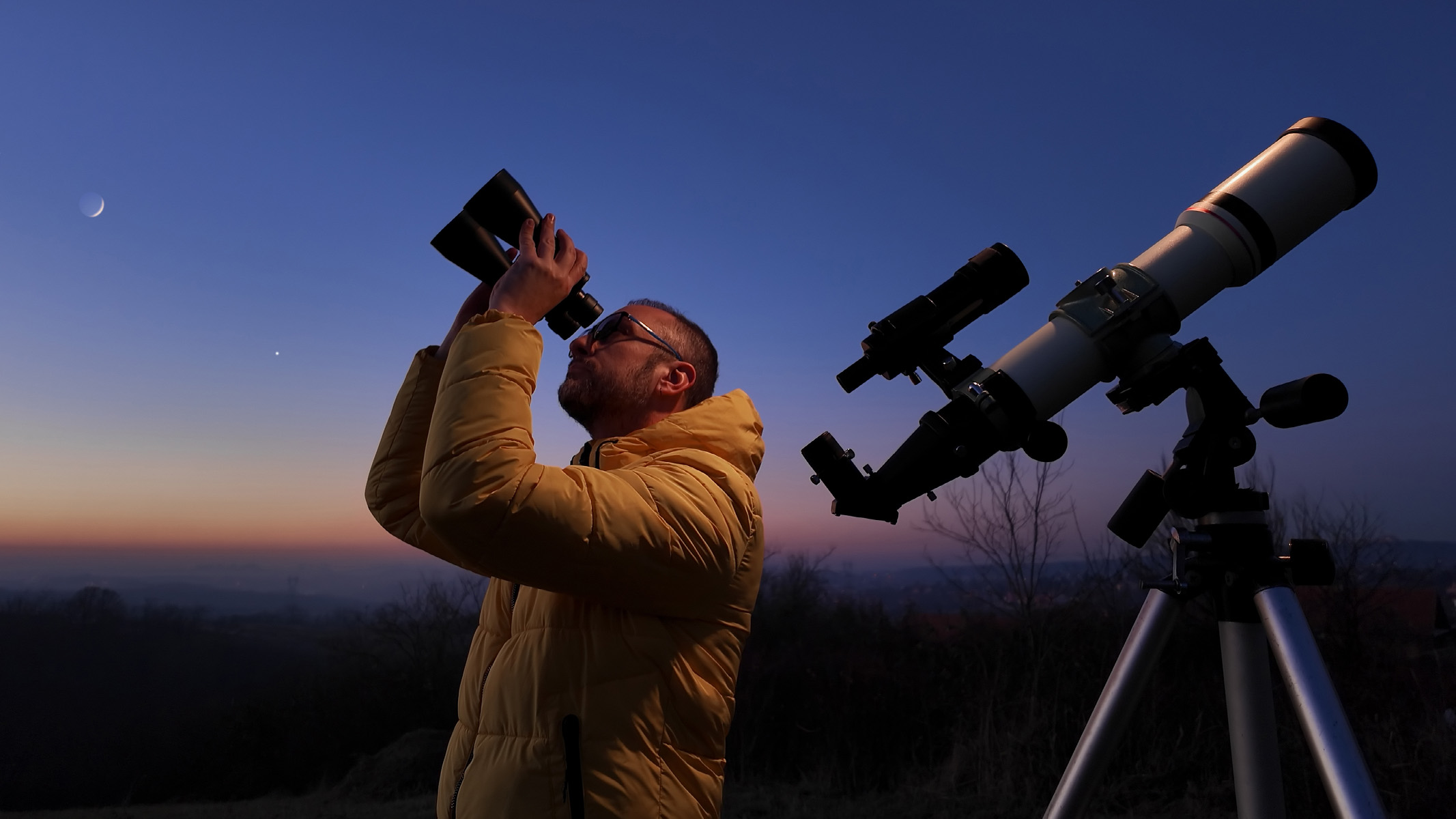 man using bioculars and a telescope at dusk