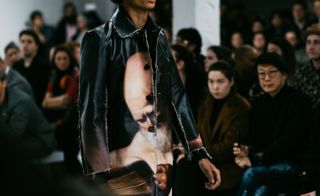 The torso of a woman wearing a black leather jacket with a mans faces printed on it walking through a crowd of seated people.