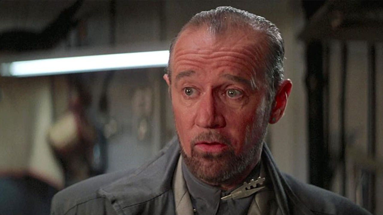 George Carlin wearing his iconic trench coat and slicked back hair in Bill and Ted's Excellent Adventure.