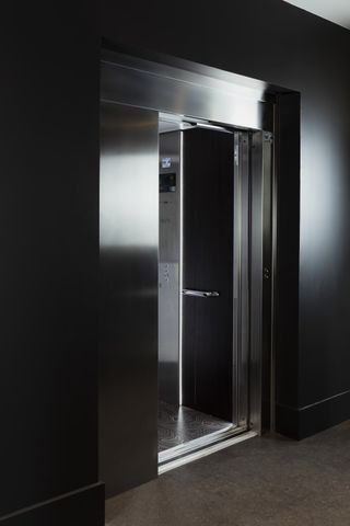 View of a space with black walls, brown floors and an Otis lift with metal doors. A partial view of the lift interior can be seen featuring patterned flooring, a metal panel with buttons and a dark coloured panel with a small handrail