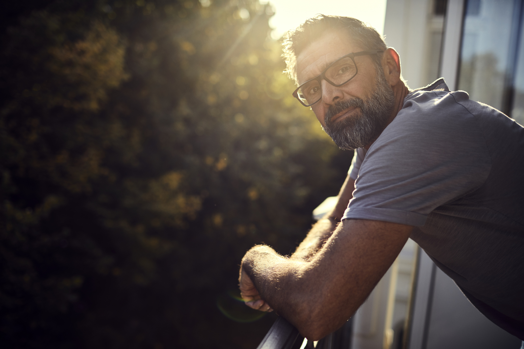 Portrait of casual mature man on balcony. The sun is behind him and his mood is thoughful or sombre. 