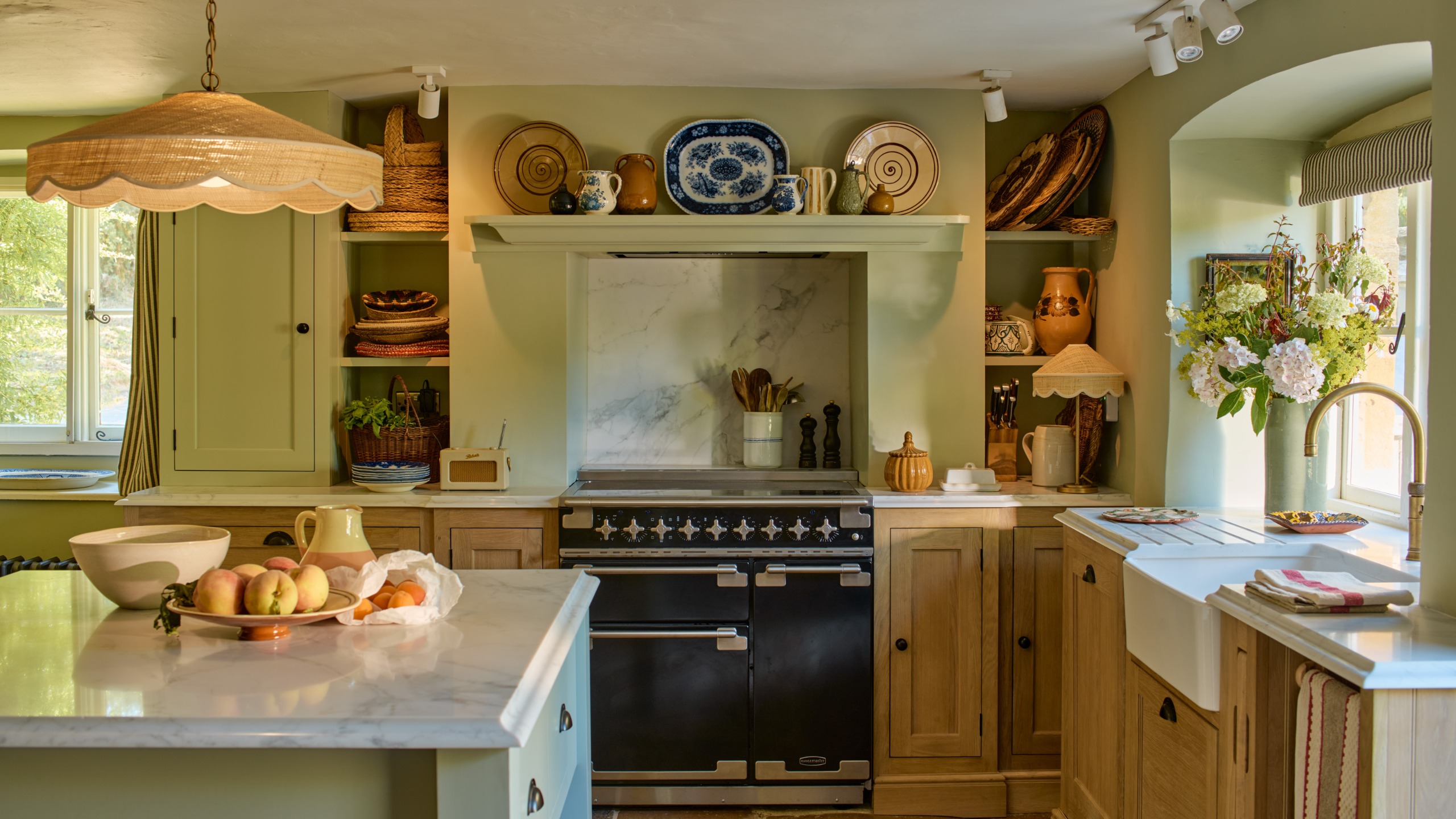 country kitchen with rattan pendant lamp above a sage island with marble worktop surrounded by oak cabinetry with black range cooker 