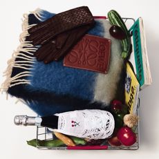 A basket that contains a loewe plaid scarf, burgundy leather gloves, a champagne bottle, a zucchini, magazines, and a red onion.