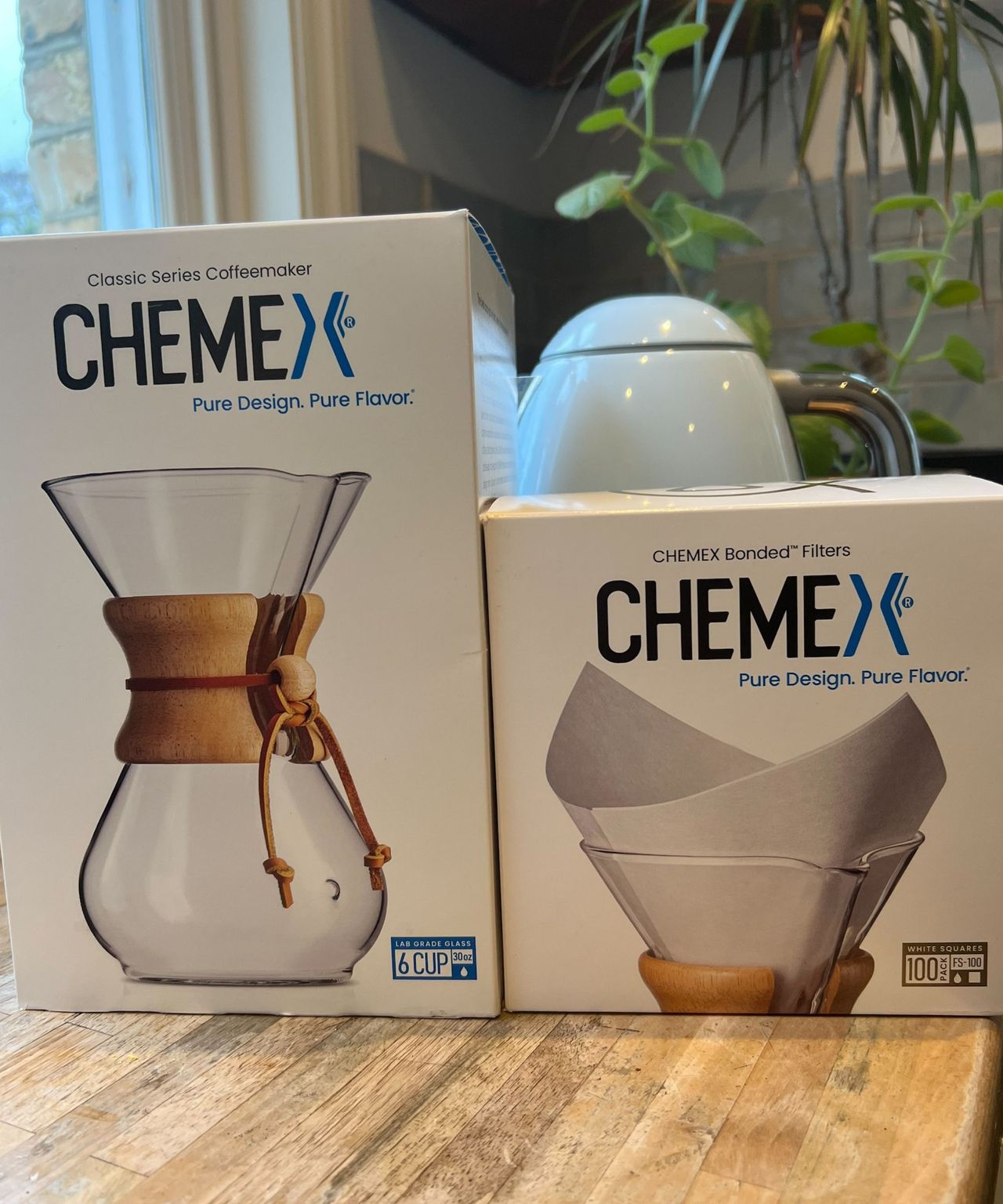 Chemex coffee maker and filters on a wooden kitchen counter with a baby blue kettle, houseplants, and a baby blue tiled backsplash behind. A window surrounded by yellow brick is seen to the left.