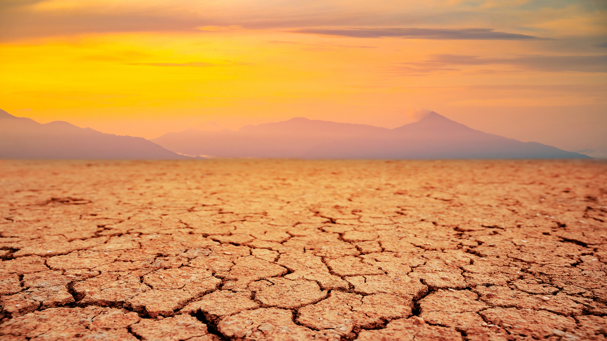 Cracked brown dirt is seen with mountains and yellow haze in the background