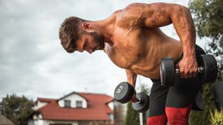 Shirtless muscular man performs bent-over row with dumbbells outside 