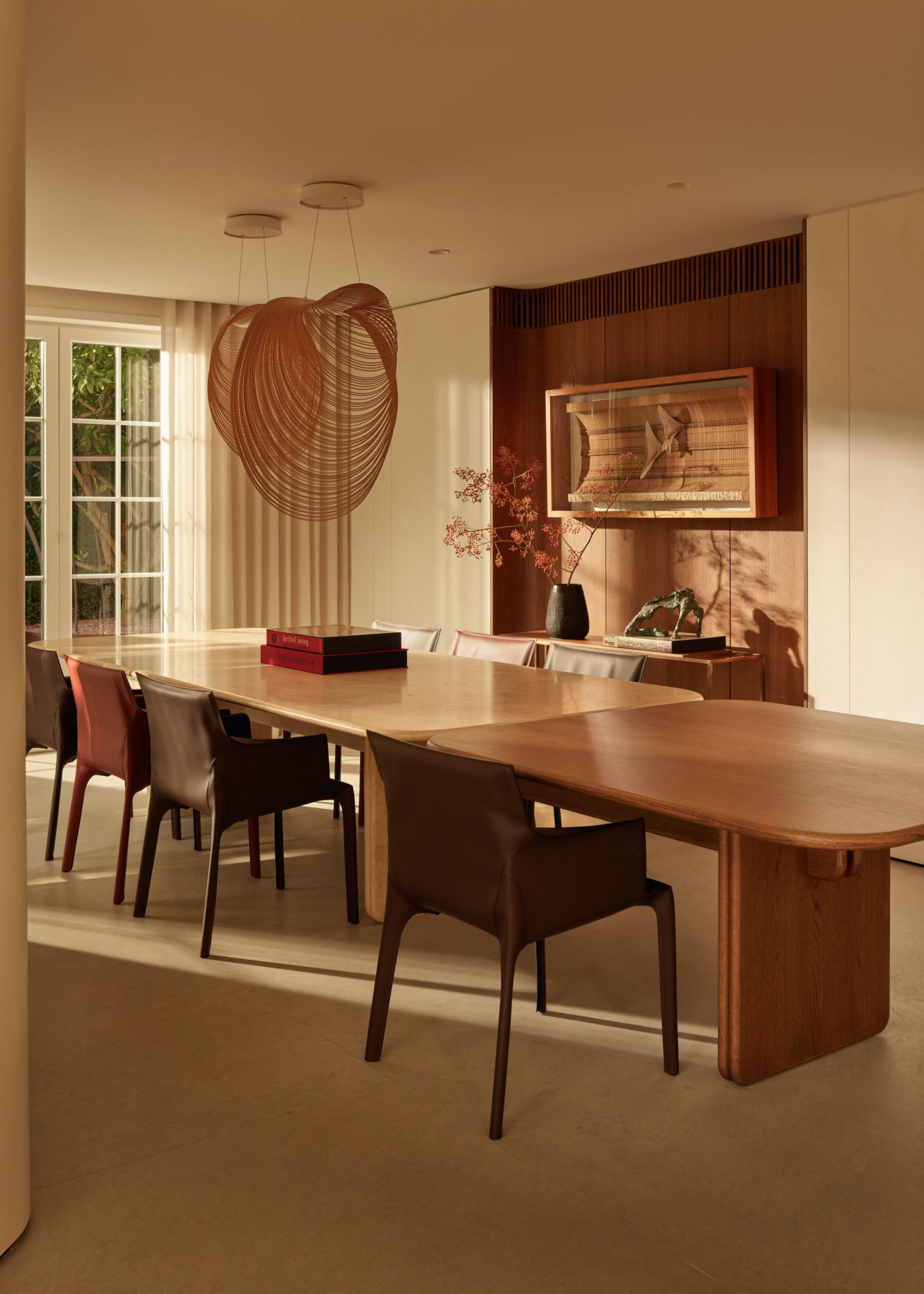 A minimalist dining room with off-white walls, textural overhead lighting, a wood dining table, leather armchairs, and a sideboard with a vase and a statue