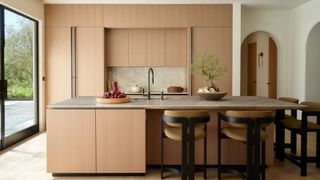 Image of a minimalist kitchen with a natural color palette and lots of light. The cabinetry is a light terracotta pink, and there is a large matching island with four yellow and black barstool chairs