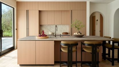 Image of a minimalist kitchen with a natural color palette and lots of light. The cabinetry is a light terracotta pink, and there is a large matching island with four yellow and black barstool chairs