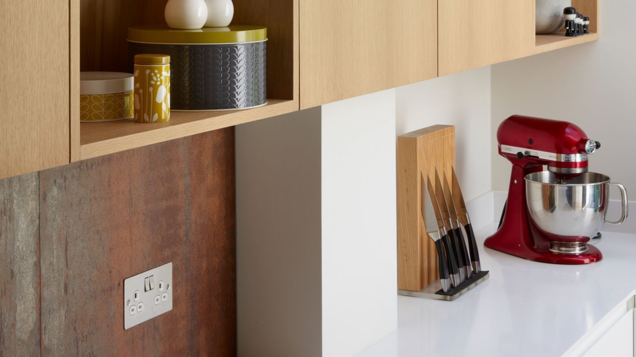 Close up of wooden kitchen wall units with kitchen jugs, cruet and tins over a white worktop with a knife block and red Kitchen Aid