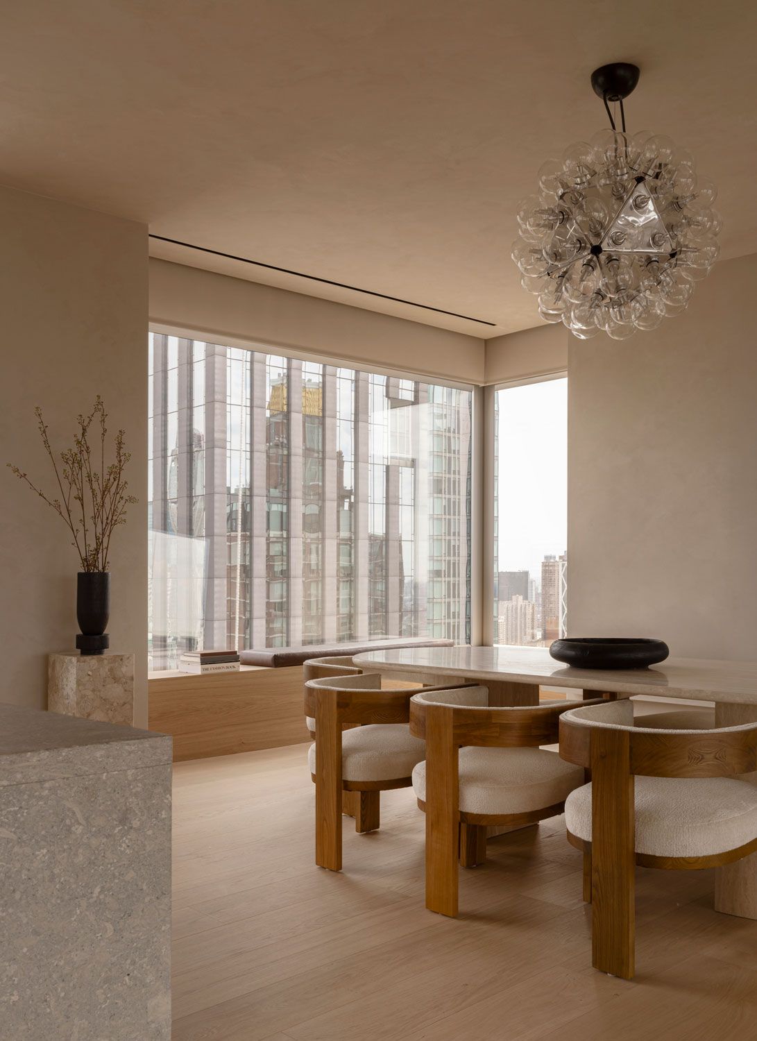 Dining area at 262 Fifth Avenue with timber chairs, a stone table, a sculptural glass pendant, and floor-to-ceiling windows overlooking Manhattan