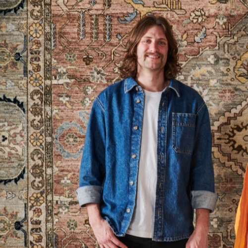 Image of a man in a white T-shirt and an open denim button down standing in front of a rug.