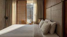 A bedroom with a double bed in the middle that has been neatly made with pillows on top of it. There are also wooden blinds installed by the window as well as curtains on each side