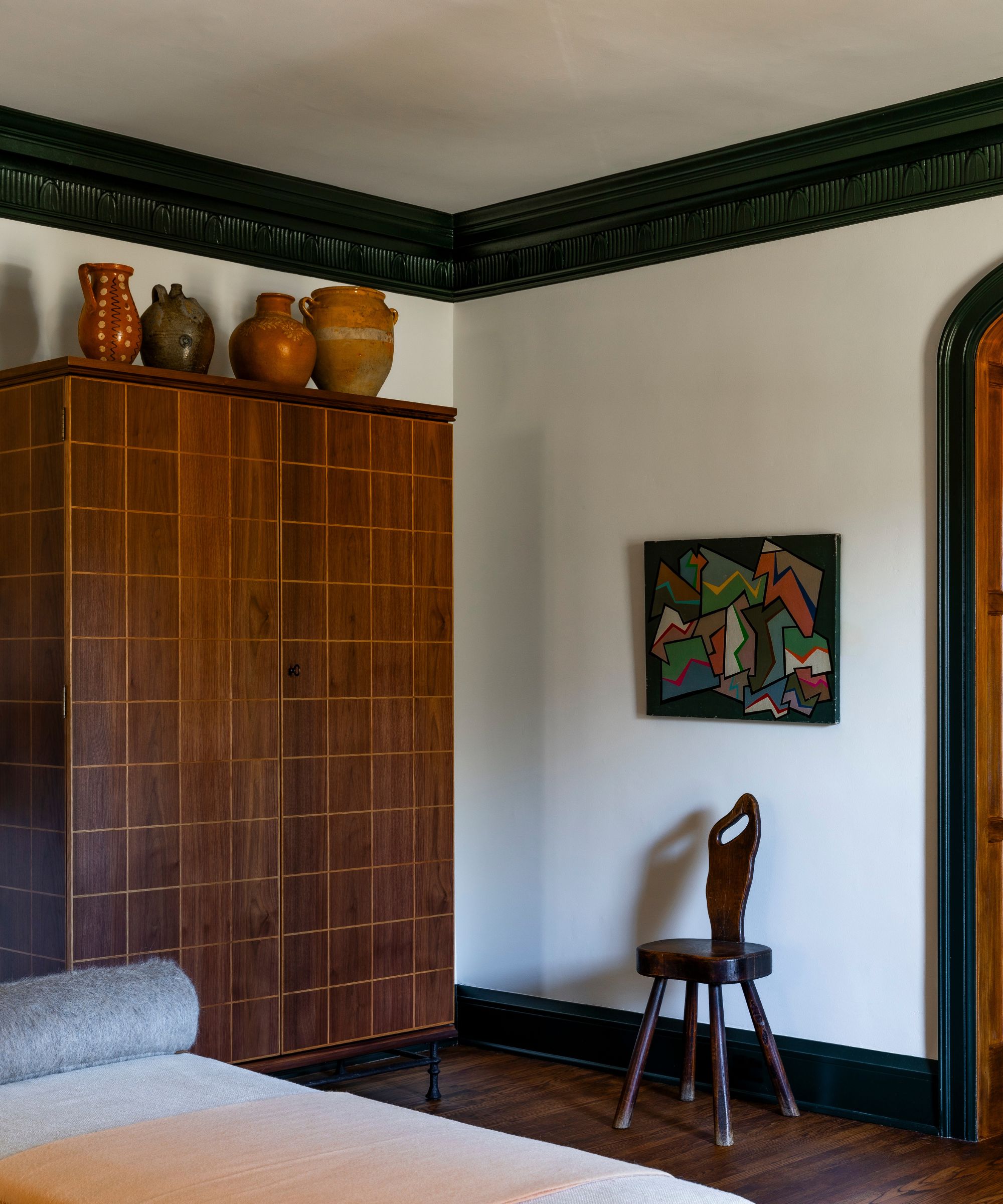 Corner of a living room with dark green ceiling trim, a vintage wardrobe with ceramic pots lined up on top, a rustic spindle chair, an abstract painting above, and a chaise lounge.