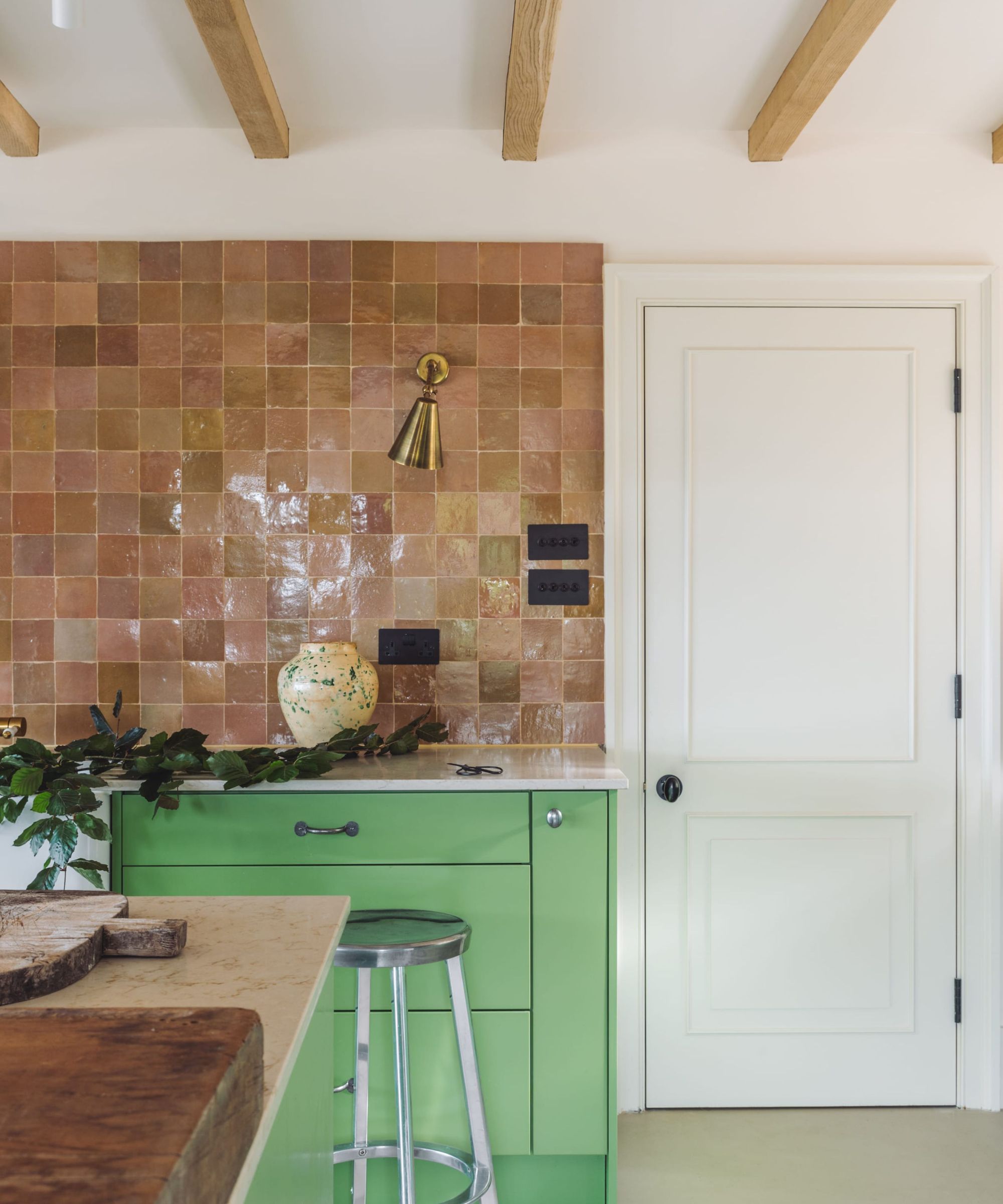 A modern kitchen with soft white walls, plaster pink zellige tiles, and pistachio green lower cabinets. Brass hardware and light wood ceiling beams.
