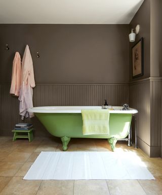 colors that go with brown, bathroom with brown painted walls, apple green roll top bath tub, tiled floor, hanging robes, artwork, stool, bathmat