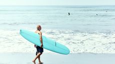 An older man carries a surfboard on the beach.