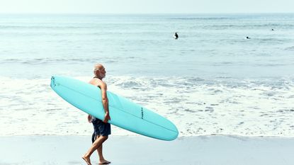 An older man carries a surfboard on the beach.