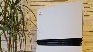 The PS5 Pro console on a wooden desk in front of a white brick background and next to a potted plant