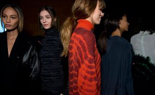 Four models - one in a black coat, one in a black and grey striped outfit, one in a red sweater, and one in dark green