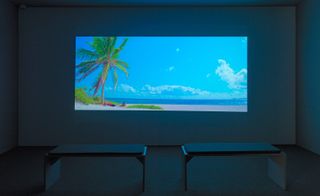 White darkened room, grey floor, two stool seats centre, projection screen on the wall of a beach, blue sky, sand, man lay under a palm tree, grass verge, blue sea