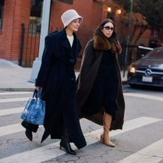 Two women are walking on a NYC cross walk. One is wearing a long black coat, bucket hat, black boots, and a blue denim handbag. The other woman is wearing a long black coat with faux fur trim on the collar and tall tan wedged boots. 