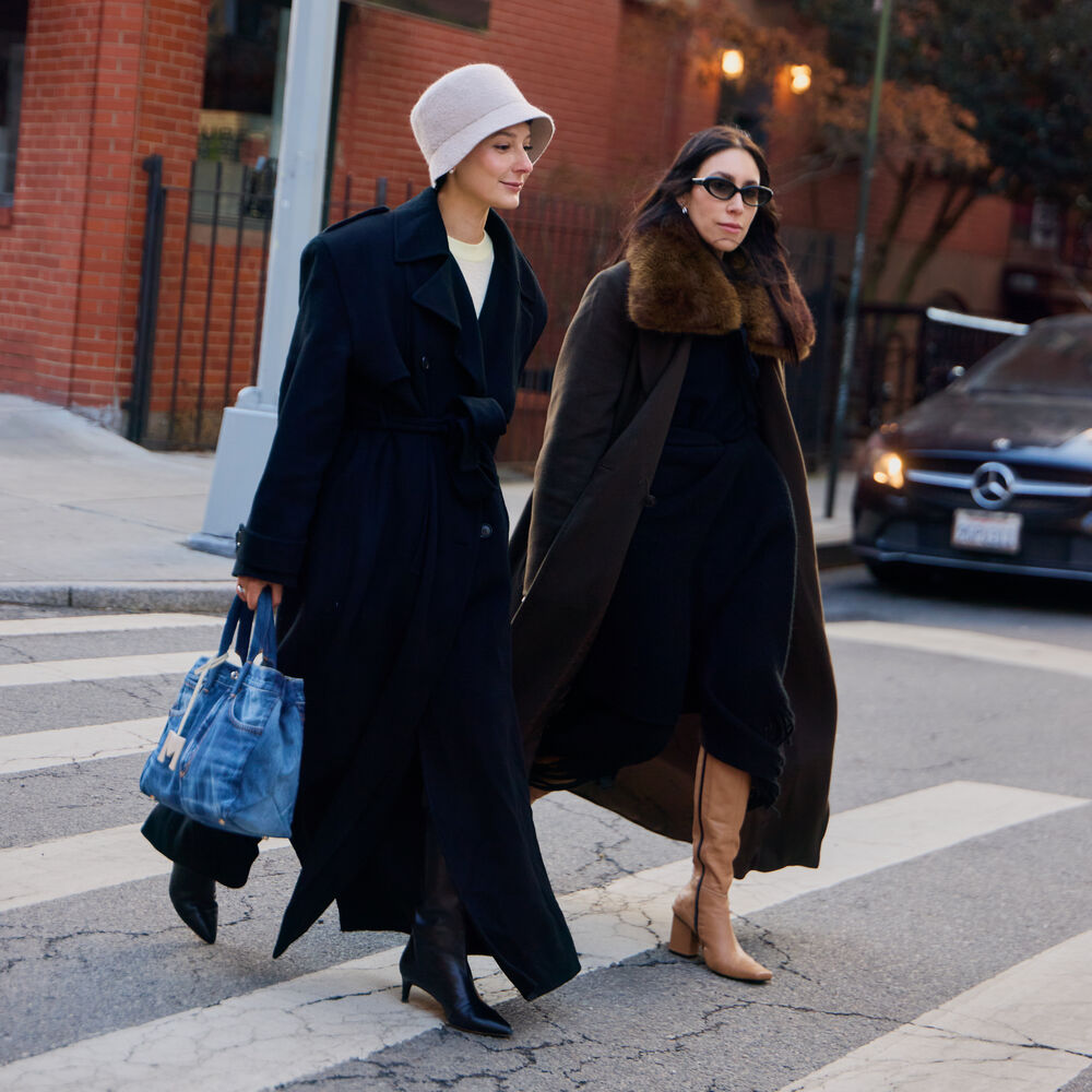 Two women are walking on a NYC cross walk. One is wearing a long black coat, bucket hat, black boots, and a blue denim handbag. The other woman is wearing a long black coat with faux fur trim on the collar and tall tan wedged boots. 